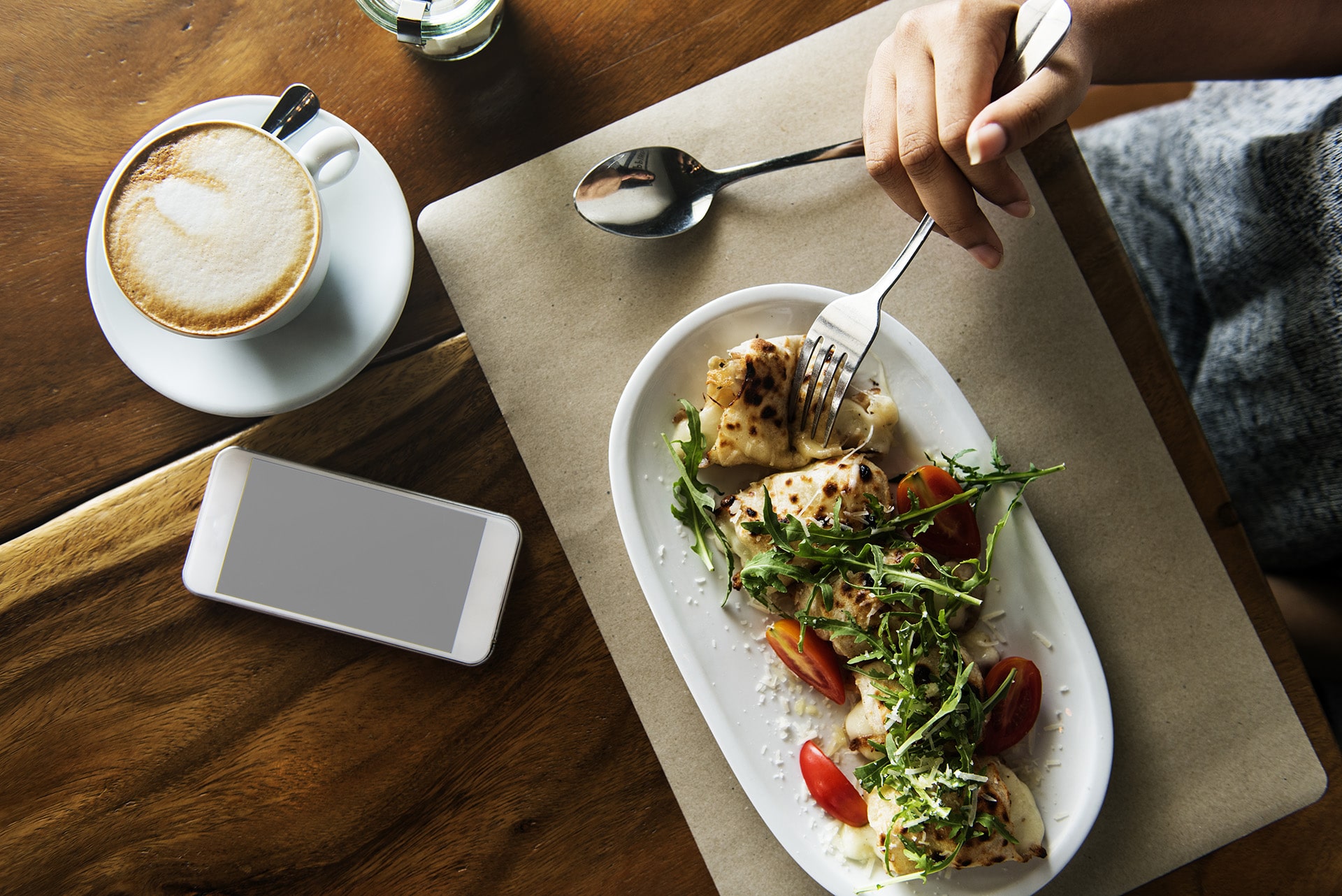A table setting featuring a plate of fresh wraps and salad, a cup of cappuccino, and a smartphone, symbolizing a casual luncheon meeting.